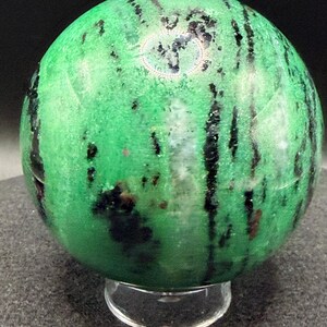 May include: A polished green sphere with black veining and dark spots, resting on a clear acrylic stand. The gemstone has a smooth, reflective surface, showcasing natural patterns and color variations.