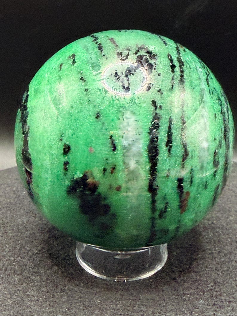 May include: A polished green sphere with black veining and dark spots, resting on a clear acrylic stand. The gemstone has a smooth, reflective surface, showcasing natural patterns and color variations.