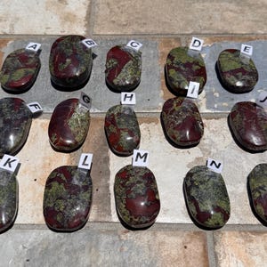 Puede incluir: Una variedad de piedras de sangre de drag&oacute;n pulidas, cada una con un patr&oacute;n &uacute;nico de tonos rojos y verdes intensos. Las piedras, de forma ovalada, est&aacute;n etiquetadas de la A a la O y miden en cent&iacute;metros.