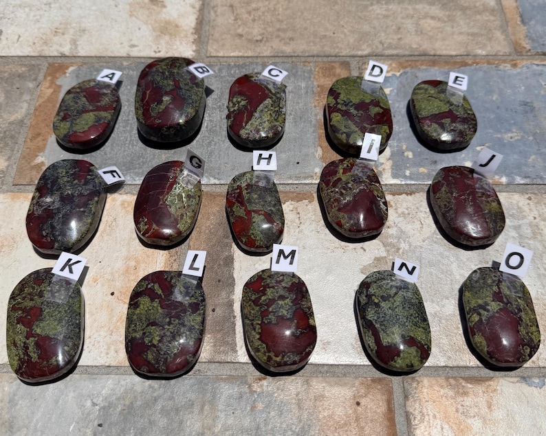 Puede incluir: Una variedad de piedras de sangre de drag&oacute;n pulidas, cada una con un patr&oacute;n &uacute;nico de tonos rojos y verdes intensos. Las piedras, de forma ovalada, est&aacute;n etiquetadas de la A a la O y miden en cent&iacute;metros.