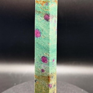 May include: A tall, rectangular crystal obelisk with a pointed top. The stone features a mottled green base color with vibrant pink and red inclusions. The obelisk is a decorative item, likely for display or spiritual use.