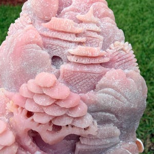 May include: A pink stone carving featuring a detailed landscape scene with tiered pagoda structures, rolling hills, and a stylized tree with layered foliage. The surface has a matte, textured finish with intricate relief work throughout the piece.
