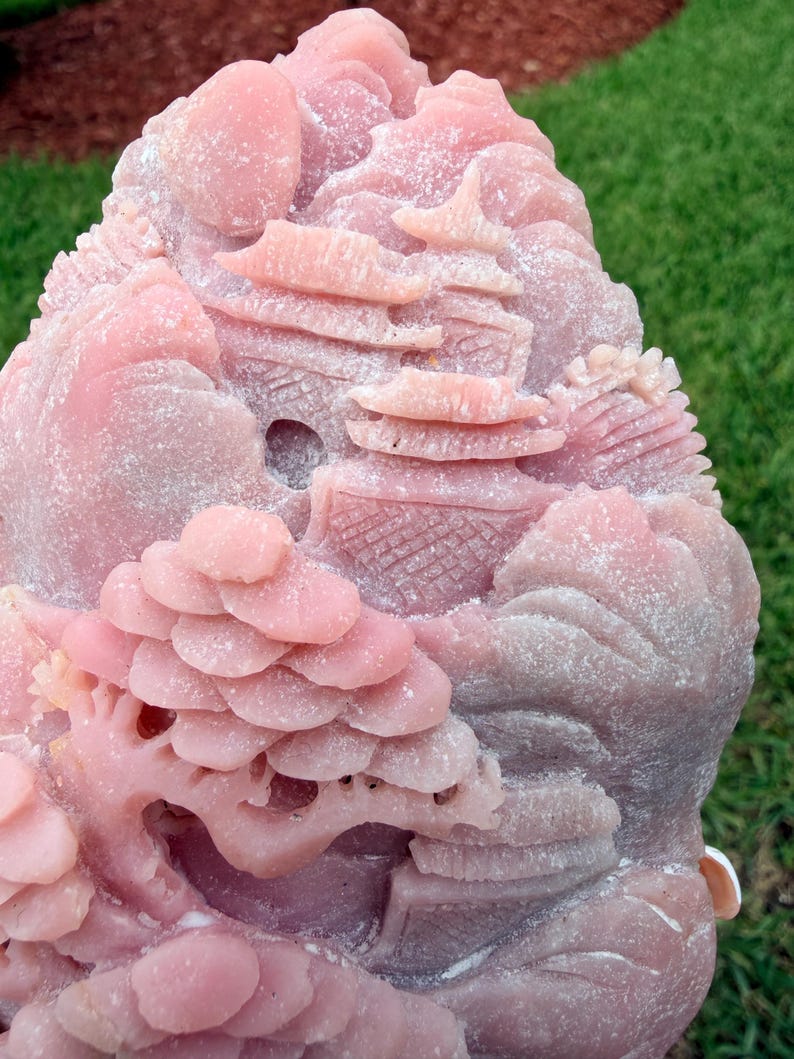 May include: A pink stone carving featuring a detailed landscape scene with tiered pagoda structures, rolling hills, and a stylized tree with layered foliage. The surface has a matte, textured finish with intricate relief work throughout the piece.