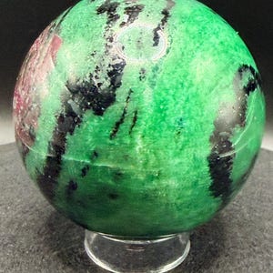 May include: A polished gemstone sphere, predominantly green with black and red veining. The sphere is displayed on a clear, circular stand, showcasing its natural patterns and textures. The gemstone is a decorative item.