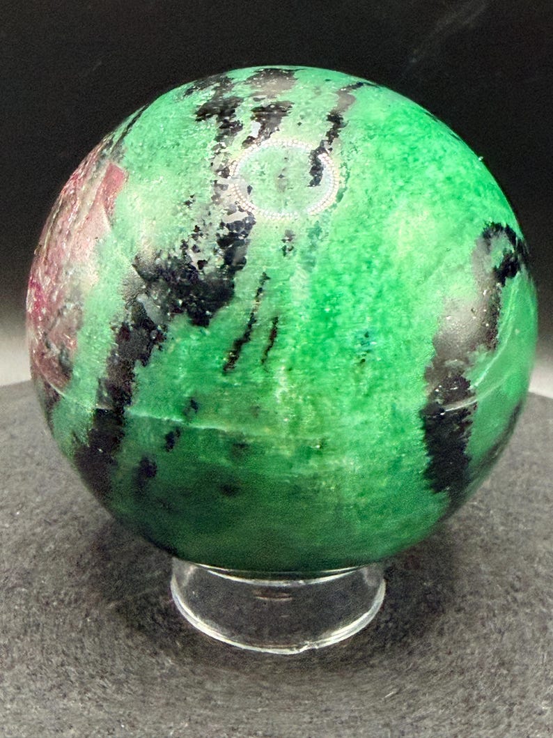May include: A polished gemstone sphere, predominantly green with black and red veining. The sphere is displayed on a clear, circular stand, showcasing its natural patterns and textures. The gemstone is a decorative item.