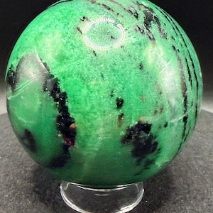 May include: A green sphere with black and dark red inclusions, resting on a clear, circular stand. The sphere has a polished surface and a marbled appearance. The overall effect is a decorative, natural stone object.