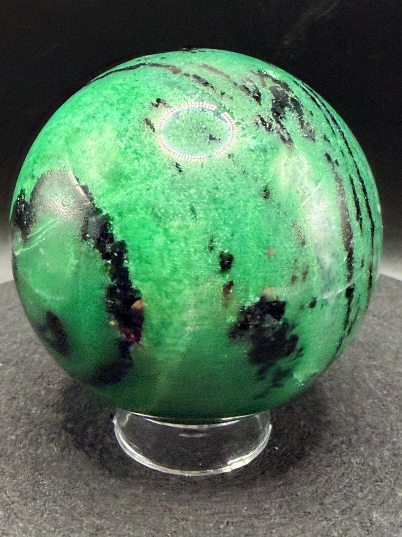 May include: A green sphere with black and dark red inclusions, resting on a clear, circular stand. The sphere has a polished surface and a marbled appearance. The overall effect is a decorative, natural stone object.