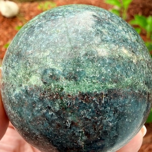 May include: A polished spherical stone featuring a mottled texture with deep forest green, teal, and dark brown mineral inclusions. The surface displays a natural crystalline pattern with varying shades of blue-green and dark earthy tones throughout.
