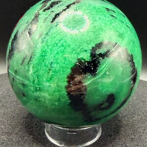 May include: A polished green sphere with black veining, resting on a clear acrylic stand. The sphere's surface has a mottled appearance, with varying shades of green and dark, irregular patterns. The overall effect is a natural, decorative object.