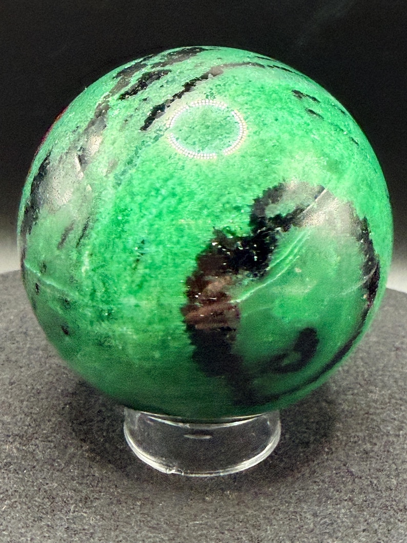 May include: A polished green sphere with black veining, resting on a clear acrylic stand. The sphere's surface has a mottled appearance, with varying shades of green and dark, irregular patterns. The overall effect is a natural, decorative object.