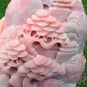 May include: A pink stone carving featuring a detailed landscape scene with layered trees, architectural structures, and rolling hills. The surface has a matte, textured finish with intricate relief work depicting a natural environment with buildings.