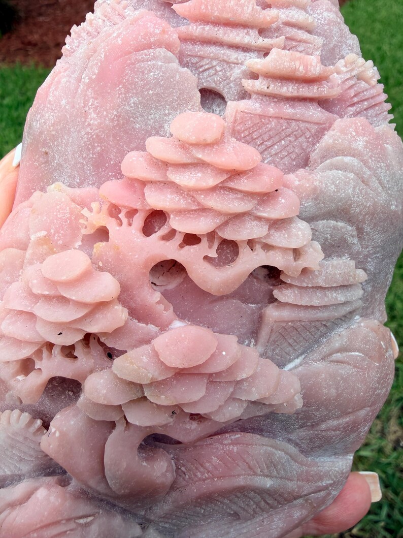 May include: A pink stone carving featuring a detailed landscape scene with layered trees, architectural structures, and rolling hills. The surface has a matte, textured finish with intricate relief work depicting a natural environment with buildings.