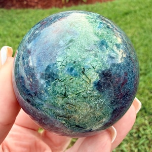 May include: A polished spherical gemstone featuring a blend of deep blue, teal, and forest green hues. The surface displays intricate internal crystalline structures, including dark needle-like inclusions and translucent patches that catch the light.
