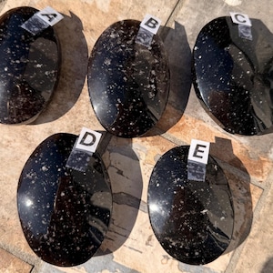 May include: Five polished, oval-shaped black stones with white speckles are arranged in a group. Each stone has a label with a letter from A to E. The stones are displayed on a light-colored surface.