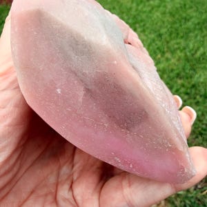 May include: A large, polished, semi-translucent pink crystal specimen with a smooth, rounded, and elongated shape. The stone features varying shades of soft pink, ranging from pale to deep rose, with subtle greyish inclusions visible within the structure.