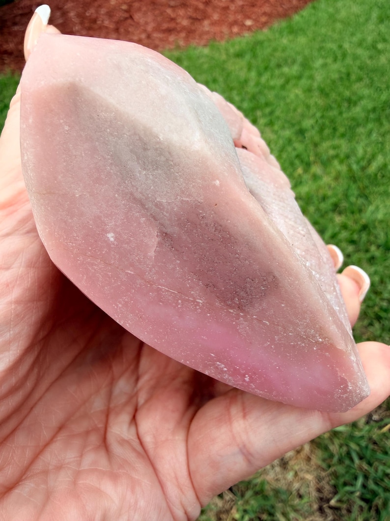 May include: A large, polished, semi-translucent pink crystal specimen with a smooth, rounded, and elongated shape. The stone features varying shades of soft pink, ranging from pale to deep rose, with subtle greyish inclusions visible within the structure.