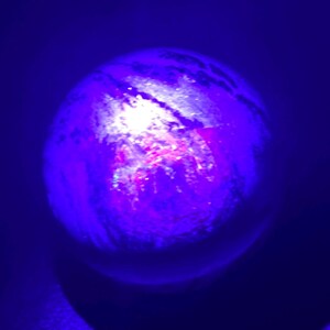 May include: A glowing, spherical object with a marbled texture, illuminated by a bright light source. The object's surface displays shades of purple, pink, and white, set against a dark blue background.