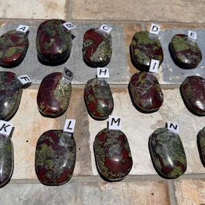 Puede incluir: Quince piedras pulidas con una mezcla de colores rojo, verde y negro. Cada piedra tiene un patr&oacute;n &uacute;nico y est&aacute; etiquetada con una letra de la A a la O en una peque&ntilde;a etiqueta blanca. Las piedras, de forma ovalada, parecen ser gemas.