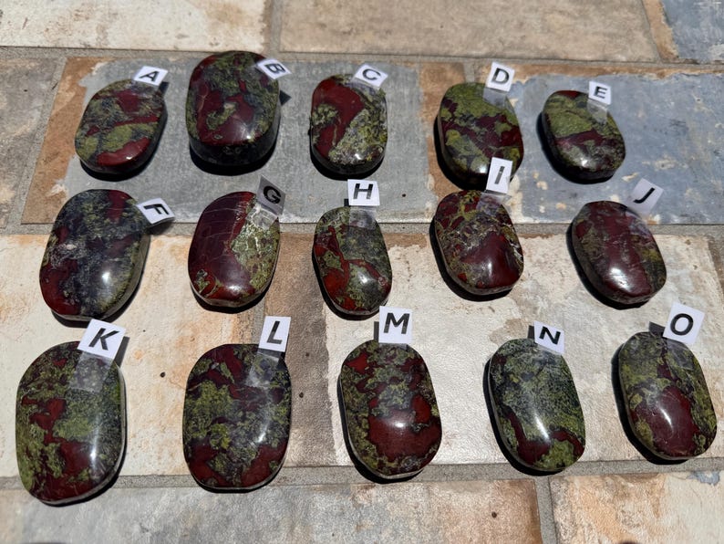 Puede incluir: Quince piedras pulidas con una mezcla de colores rojo, verde y negro. Cada piedra tiene un patr&oacute;n &uacute;nico y est&aacute; etiquetada con una letra de la A a la O en una peque&ntilde;a etiqueta blanca. Las piedras, de forma ovalada, parecen ser gemas.
