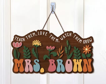 Pode incluir: Uma placa de madeira marrom com o texto "Teach them, love them, watch them grow" e "Mrs. Brown" em uma fonte colorida e caprichosa. A placa é decorada com flores e vegetação coloridas.