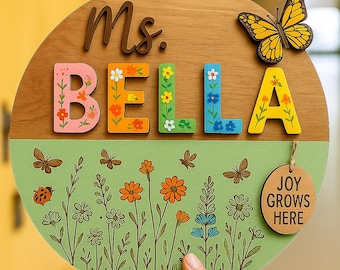 May include: A wooden round sign with the name "Ms. BELLA" in colourful, floral-decorated letters. A yellow butterfly and a "Joy Grows Here" tag are attached. The bottom half features a green field with drawn flowers and butterflies.