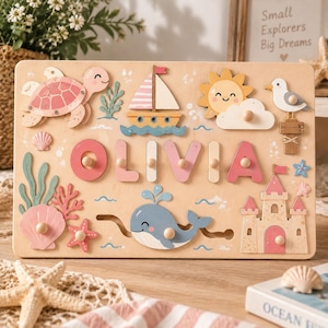 May include: A rectangular wooden name puzzle featuring the name OLIVIA in pink and cream letters with knobs. The board displays nautical illustrations including a pink turtle, a sailboat, a smiling sun, a seagull, a whale, and a sandcastle.