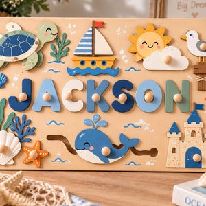 May include: A wooden name puzzle featuring the name JACKSON in blue and green letters with knobs. The board displays nautical illustrations including a sea turtle, sailboat, sun, seagull, whale, and sandcastle with various sea life accents.