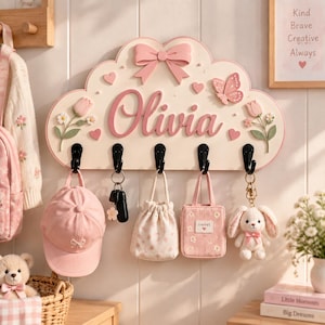 Könnte beinhalten: Eine wolkenförmige Wandgarderobe in Creme mit rosa Schleife, Schmetterling und Tulpen. Der Name Olivia ist in rosa Schreibschrift zu sehen. Sie hat fünf schwarze Haken, an denen eine rosa Baseballkappe, zwei kleine Taschen und ein Hasenanhänger hängen.