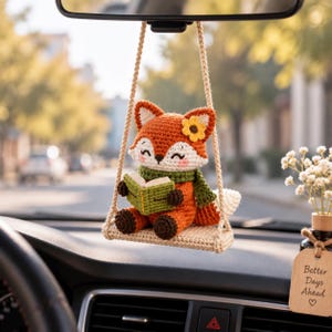 May include: A crocheted fox ornament hanging from a rearview mirror, sitting on a small swing. The orange and white fox wears a green scarf and a yellow flower, holding a green book. A wooden tag nearby reads Better Days Ahead with a heart.