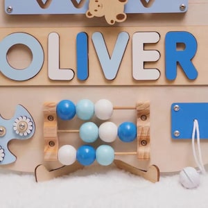 May include: A wooden activity board featuring the name OLIVER in blue and white letters. The board includes a small wooden bear, a blue spinner with white flower accents, a three-row abacus with blue and white beads, and a blue pull-cord mechanism.