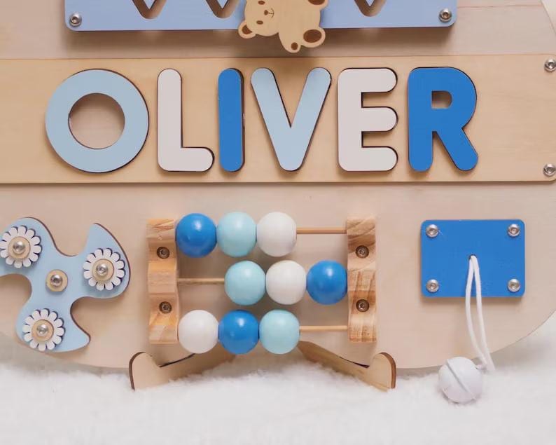 May include: A wooden activity board featuring the name OLIVER in blue and white letters. The board includes a small wooden bear, a blue spinner with white flower accents, a three-row abacus with blue and white beads, and a blue pull-cord mechanism.