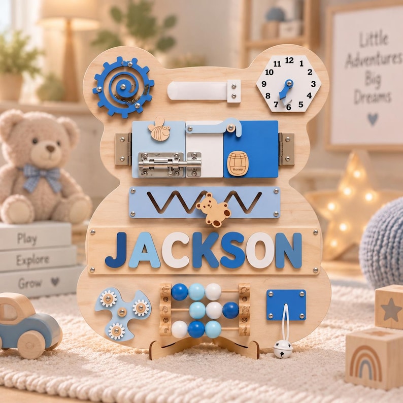 May include: A bear-shaped wooden activity board featuring the name JACKSON in blue letters. It includes a blue gear, a clock, a metal latch, a honey barrel icon, a sliding bear piece, a bead abacus, and a white bell on a string for sensory play.
