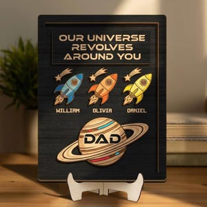 May include: A wooden plaque with the text "OUR UNIVERSE REVOLVES AROUND YOU". It features three colorful rockets and a planet with the word "DAD". The names William, Olivia, and Daniel are below the rockets.