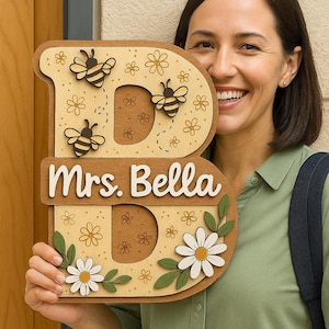 May include: Wooden letter "B" shaped sign with a light brown border, featuring bees, flowers, and the name "Mrs. Bella". The sign is decorated with daisies and other floral elements, perfect for a home or classroom.
