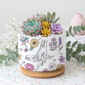 May include: A white ceramic planter with a wooden base, filled with colorful succulents. The planter is decorated with floral designs and the names "Amy", "Sarah", "Jason", and "Mama".