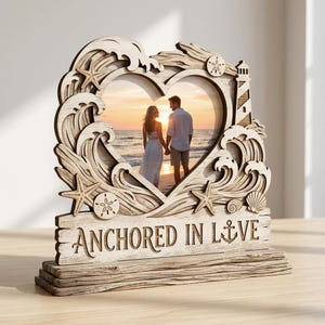 Puede incluir: Marco de fotos de madera en forma de corazón, con temática de playa. El marco presenta olas, estrellas de mar y un faro cortados con láser. Las palabras "ANCHORED IN LOVE" están talladas en la base. Contiene una foto de una pareja.