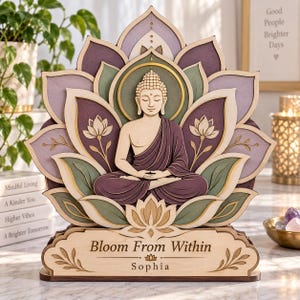 Puede incluir: Un expositor decorativo de madera en capas con una figura central de Buda sentado en posición de loto, rodeado de pétalos de loto morados y verdes. La base incluye el texto grabado "Bloom From Within" y el nombre "Sophia" en negro.