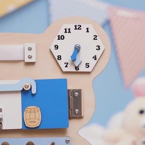 May include: A wooden activity board featuring a hexagonal clock with black numbers and blue hands, a white strap, a blue door with a honey barrel graphic labeled Honey, and a silver metal latch. The board is light wood with various mechanical elements.