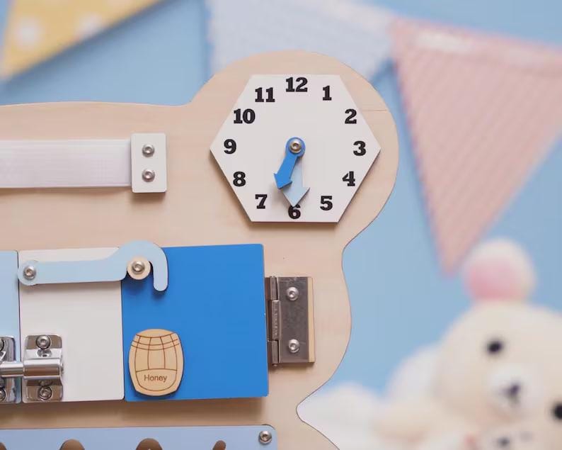 May include: A wooden activity board featuring a hexagonal clock with black numbers and blue hands, a white strap, a blue door with a honey barrel graphic labeled Honey, and a silver metal latch. The board is light wood with various mechanical elements.