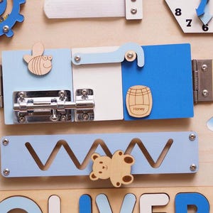 May include: A wooden sensory activity board featuring a blue gear with beads, a clock face, and a sliding metal bolt latch on light blue, white, and blue panels. A wooden bee, a honey barrel icon, and a bear-shaped slider on a zigzag track are included.