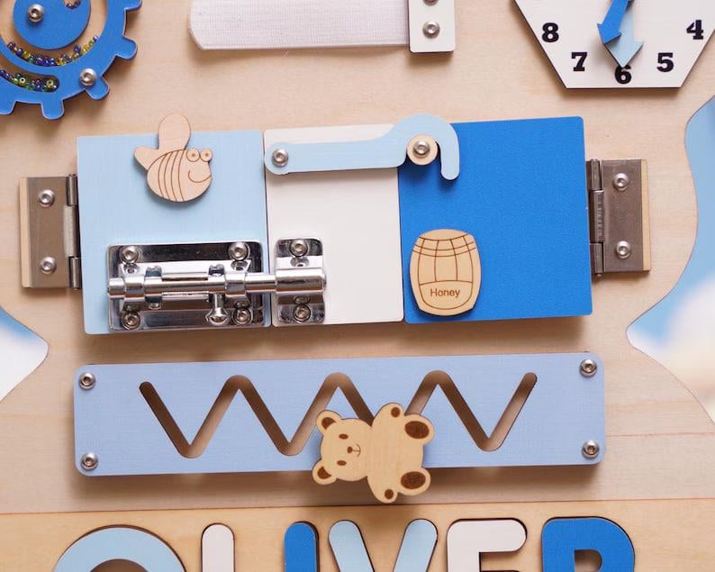 May include: A wooden sensory activity board featuring a blue gear with beads, a clock face, and a sliding metal bolt latch on light blue, white, and blue panels. A wooden bee, a honey barrel icon, and a bear-shaped slider on a zigzag track are included.