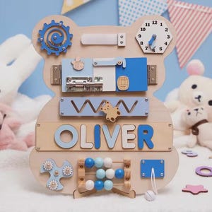 May include: A bear-shaped wooden busy board featuring a blue gear, a clock with movable hands, a metal sliding latch, a zigzag track with a bear slider, the name OLIVER in blue letters, a bead abacus, a blue spinner, and a white cord with a bead.