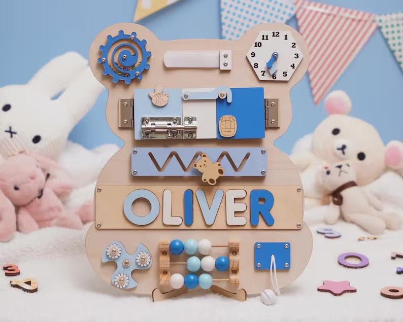 May include: A bear-shaped wooden busy board featuring a blue gear, a clock with movable hands, a metal sliding latch, a zigzag track with a bear slider, the name OLIVER in blue letters, a bead abacus, a blue spinner, and a white cord with a bead.