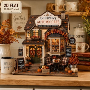 Pode incluir: Um modelo em miniatura do Anderson's Autumn Cafe com telhado castanho, portas verdes com coroas e uma janela saliente. O exterior está decorado com folhas de outono, abóboras e placas com os dizeres Coffee, Good Friends, Cozy Days e Menu.