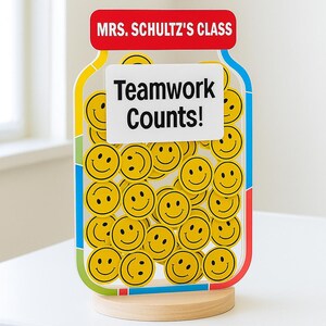 Peut inclure: Décoration de classe en forme de bocal coloré avec une base en bois. Le bocal est rempli de visages souriants jaunes et porte l'inscription "Teamwork Counts!". Le haut du bocal indique "MRS. SCHULTZ'S CLASS".
