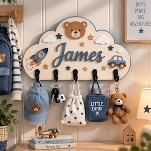 May include: A cloud-shaped wooden wall hook rack featuring the name James in blue script, a brown bear face, a rocket, a car, and stars. It has five black hooks holding a blue baseball cap, keys, a star-patterned pouch, a blue bag, and a teddy bear.