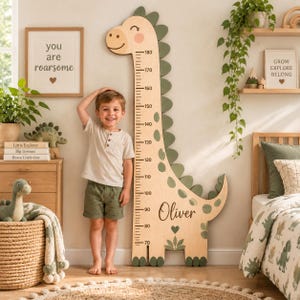 Può includere: Un metro di crescita in legno color chiaro a forma di dinosauro con una scala da 70 a 180 centimetri, decorato con accenti verdi e il nome Oliver stampato in corsivo nero, posizionato verticalmente contro una parete in una camera da letto.
