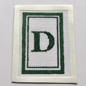 May include: A rectangular needlepoint canvas featuring a centered dark green letter D. The letter is enclosed within a double-lined dark green rectangular border. The design is stitched onto a white grid-patterned fabric with a plain white border.