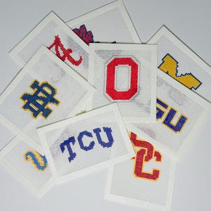 Può includere: Una collezione di tele quadrate bianche per ricamo a mezzo punto con vari loghi universitari cuciti con filo colorato, tra cui le lettere O, TCU e altri emblemi in rosso, blu e giallo su una base di tessuto con motivo a griglia.
