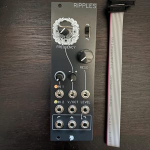 Könnte beinhalten: Schwarzes elektronisches Modul mit der Aufschrift "Ripples", Knöpfen, Buchsen und den Wörtern "Frequency" und "Reso". Ein Flachbandkabel befindet sich rechts. Das Modul hat ein dekoratives Blumendesign um den Frequenzknopf.